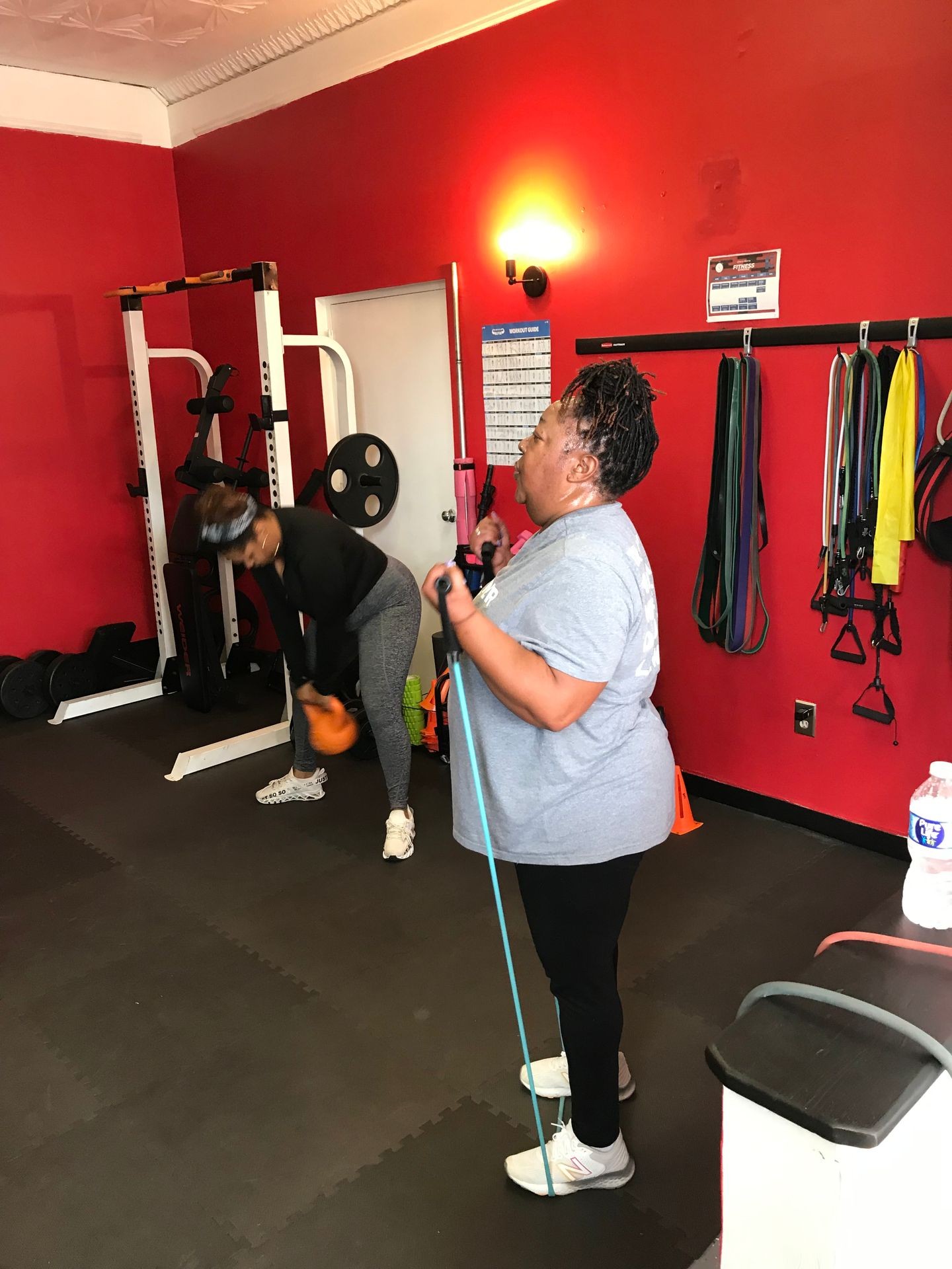 Two women exercising in a red-walled gym with fitness equipment and resistance bands.