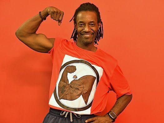 Man in a red shirt flexing his arm muscle, standing against a red background and smiling.