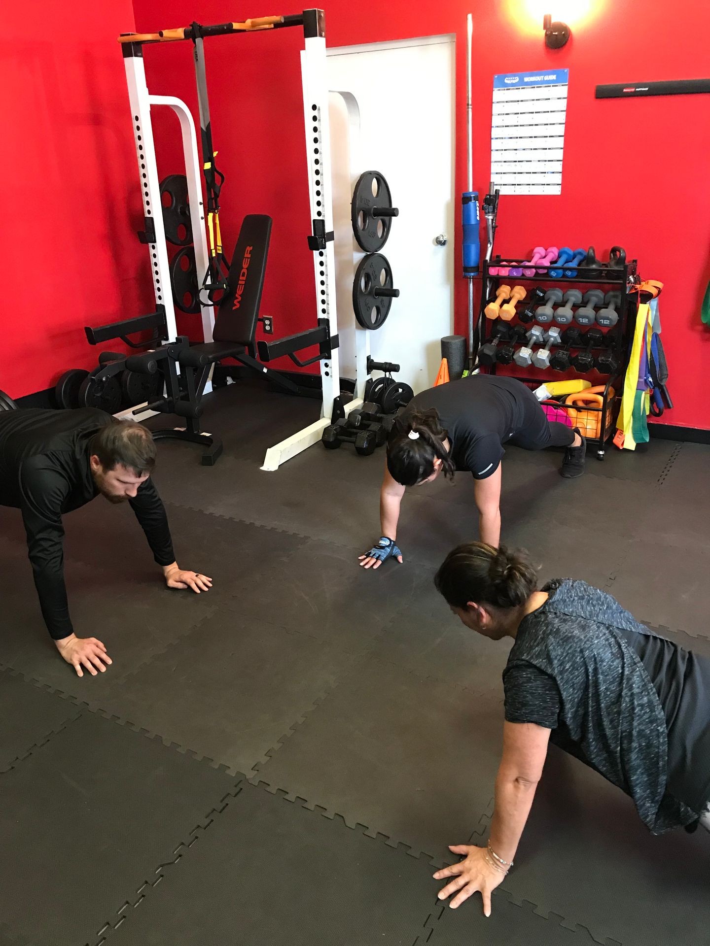 Individuals performing a plank exercise on a gym floor with various weights and fitness equipment in the background.