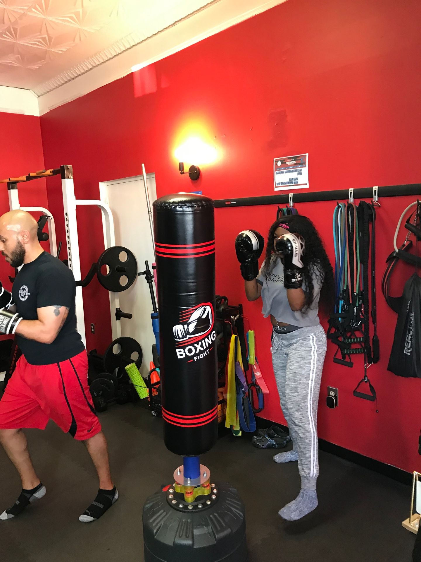 Man and woman practicing boxing with a punching bag in a brightly lit gym with red walls.